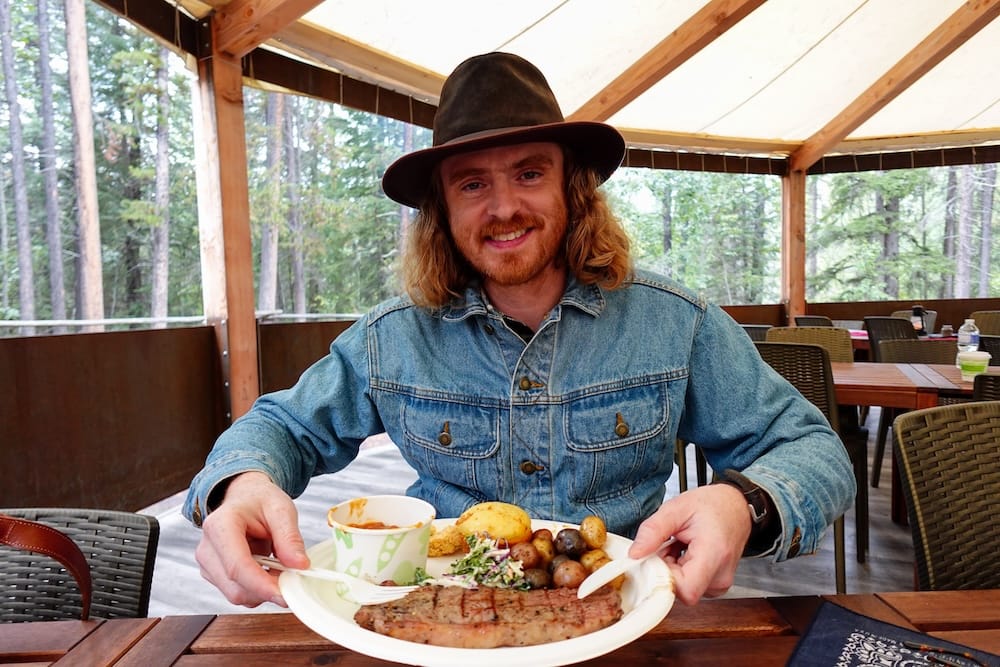 Comida BBQ con steak del Banff Cowboy Cookout en Alberta, Canadá, con Samuel Jeffery Samuel Jeffery sonríe frente a una comida BBQ con steak, papas, coleslaw y porotos en el Banff Cowboy Cookout de Alberta, Canadá, sentado bajo un refugio rústico en el bosque.