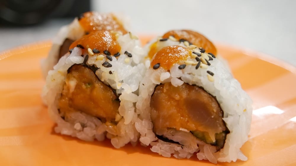 Primer plano del Cowboy Roll con carne sellada a la vista en el interior, cubierto con salsa y semillas de sésamo, sobre un plato naranja en Point Sushi en Calgary, Alberta, Canadá.