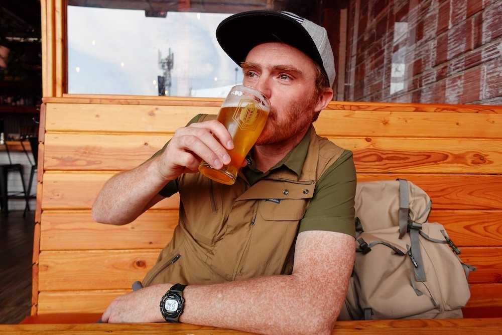 Enjoying a beer at Fire Hall Kitchen and Tap Nomadic Samuel Jeffery enjoys a chilled pint of craft beer while seated in a wooden booth at Fire Hall Kitchen and Tap in Cranbrook, BC, dressed for early autumn with a cap, vest, and hiking shirt during a relaxed break from exploring the city.