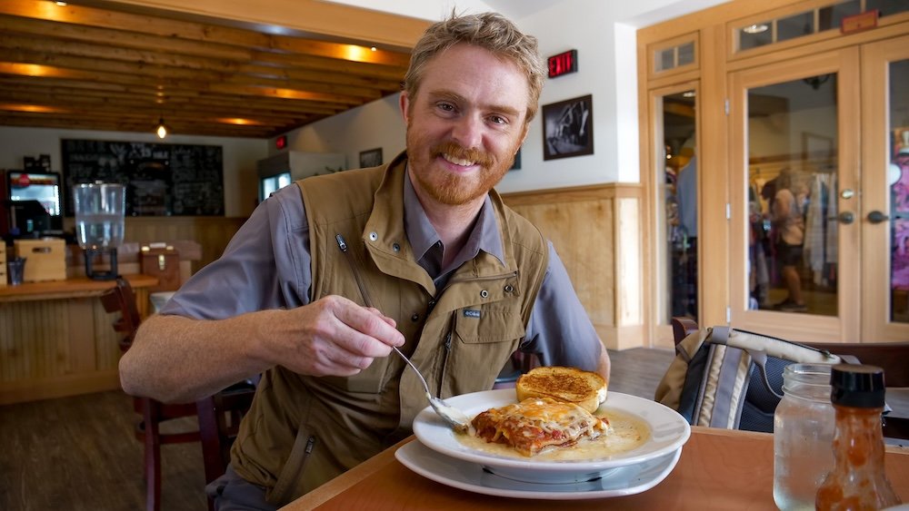 Cranbrook, British Columbia comfort food at the 19th Hole at St. Eugene Resort, with Nomadic Samuel smiling while enjoying a plate of lasagne topped with melted cheese and served with garlic bread during a relaxed resort dinner.