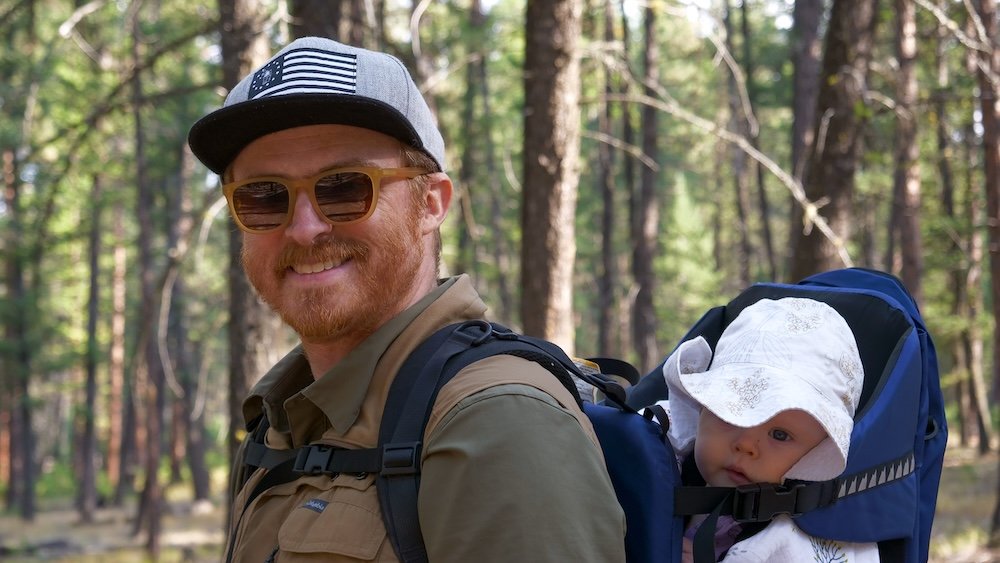 Cranbrook Community Forest in summer, BC: Nomadic Samuel hikes through sunny pine woods with baby Aurelia riding in a backpack carrier, both smiling and wide-eyed—an easy family-friendly outdoor adventure near town.