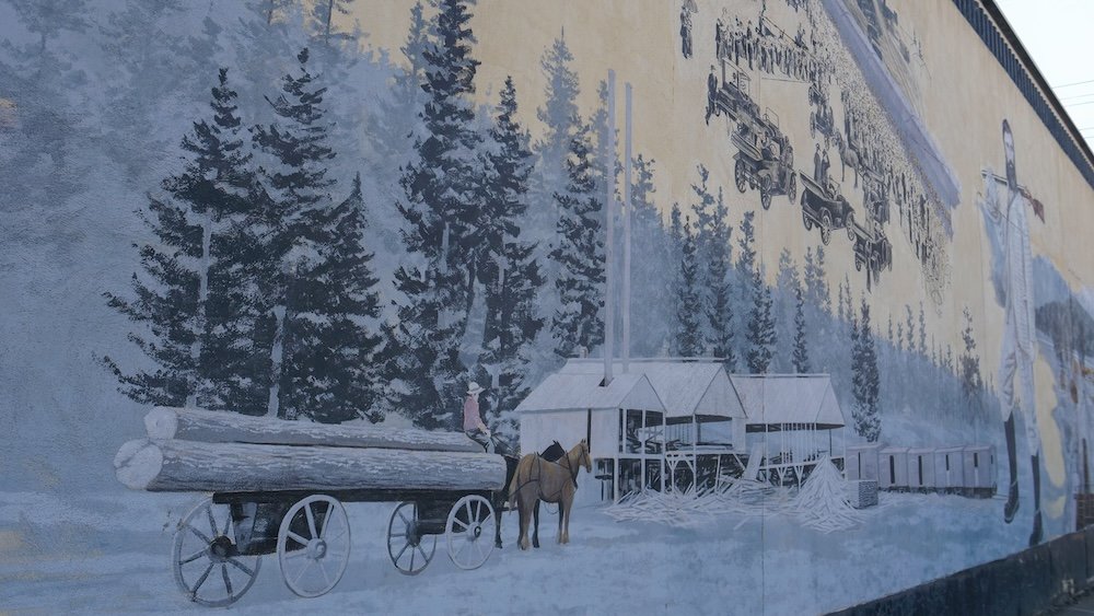 Downtown Cranbrook, BC features a large historic mural depicting early settlement scenes with horses, logging wagons, railcars, and forest landscapes, showcasing the city’s heritage through detailed public art along the streetscape.
