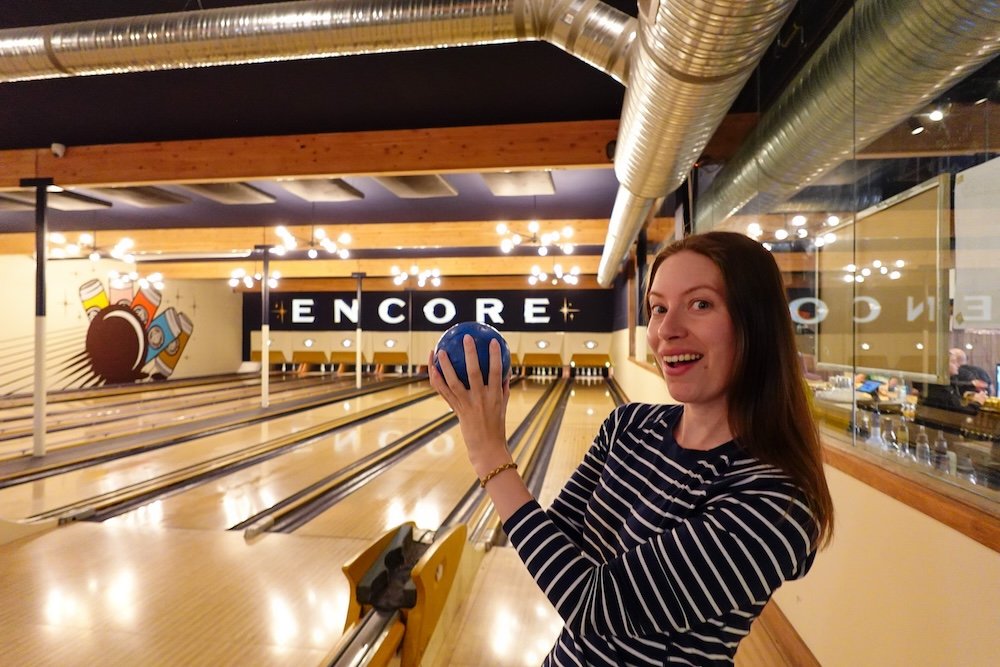 Cranbrook BC bowling lanes at Encore Brewing during a lively date night, with Audrey Bergner holding a blue five-pin bowling ball and smiling before her turn, capturing the fun, competitive energy of bowling paired with pizza and a relaxed night out.