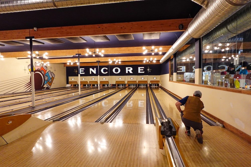 Encore Brewing in Cranbrook, British Columbia during summer, showing Nomadic Samuel bowling on vintage-style lanes inside the lively brewpub, highlighting a fun evening activity that combines craft beer, pizza, and classic bowling in the city.