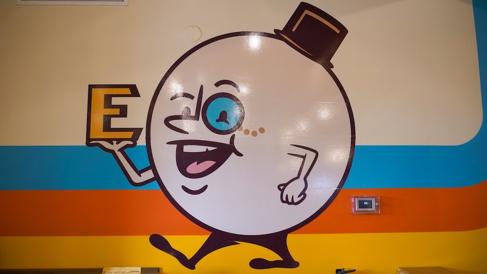 Cranbrook BC interior wall art at Encore Brewing featuring the playful bowling logo character holding an “E,” set against colorful retro stripes, highlighting the venue’s fun branding and casual entertainment vibe tied to bowling, pizza, and craft beer.