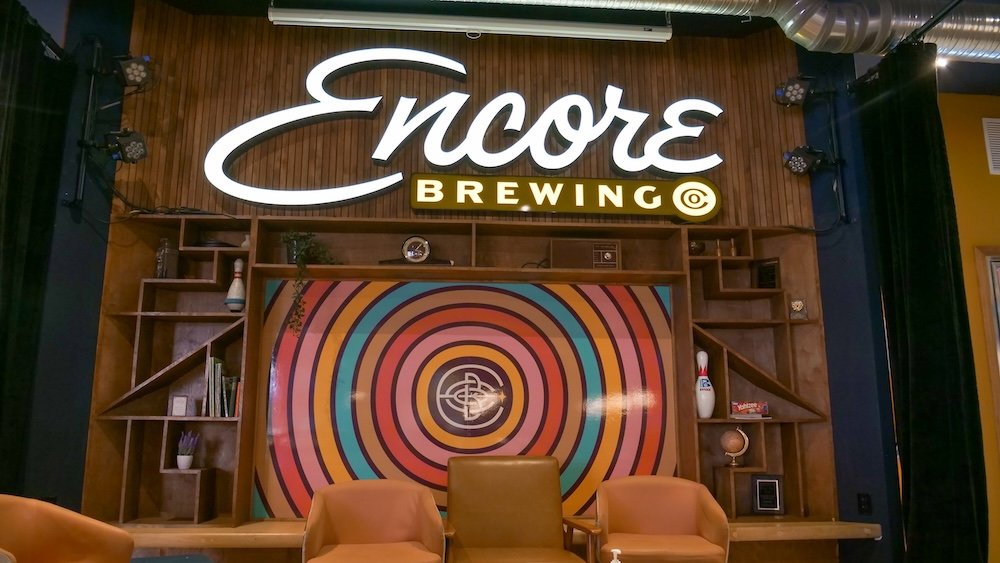Cranbrook BC interior design at Encore Brewing featuring the illuminated Encore Brewing sign, retro-inspired circular mural, and cozy seating area, highlighting the venue’s stylish mix of modern brewery aesthetics and playful design that sets the tone for bowling and pizza nights.