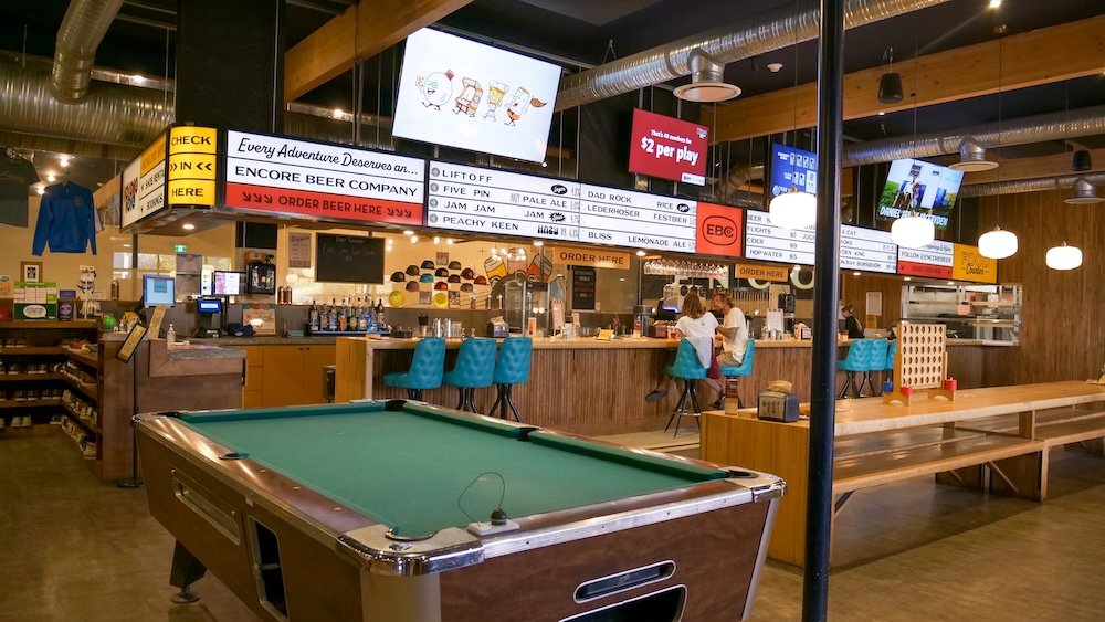 Cranbrook BC interior of Encore Brewing showing a pool table in the foreground with the bar, beer menu boards, and seating area behind it, highlighting the venue’s mix of casual games, craft beer, and relaxed social spaces beyond bowling lanes.