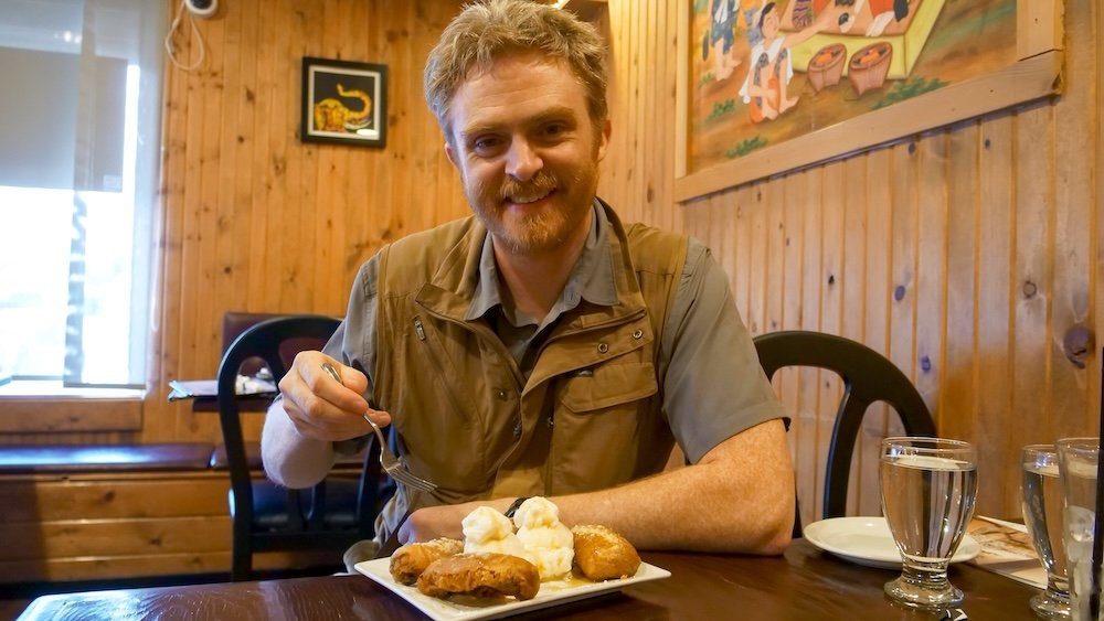 Cranbrook, British Columbia dessert at Family Thai Restaurant, with Nomadic Samuel smiling while enjoying deep-fried bananas topped with ice cream and whipped cream inside the cozy, wood-paneled dining room during a comforting Thai meal.