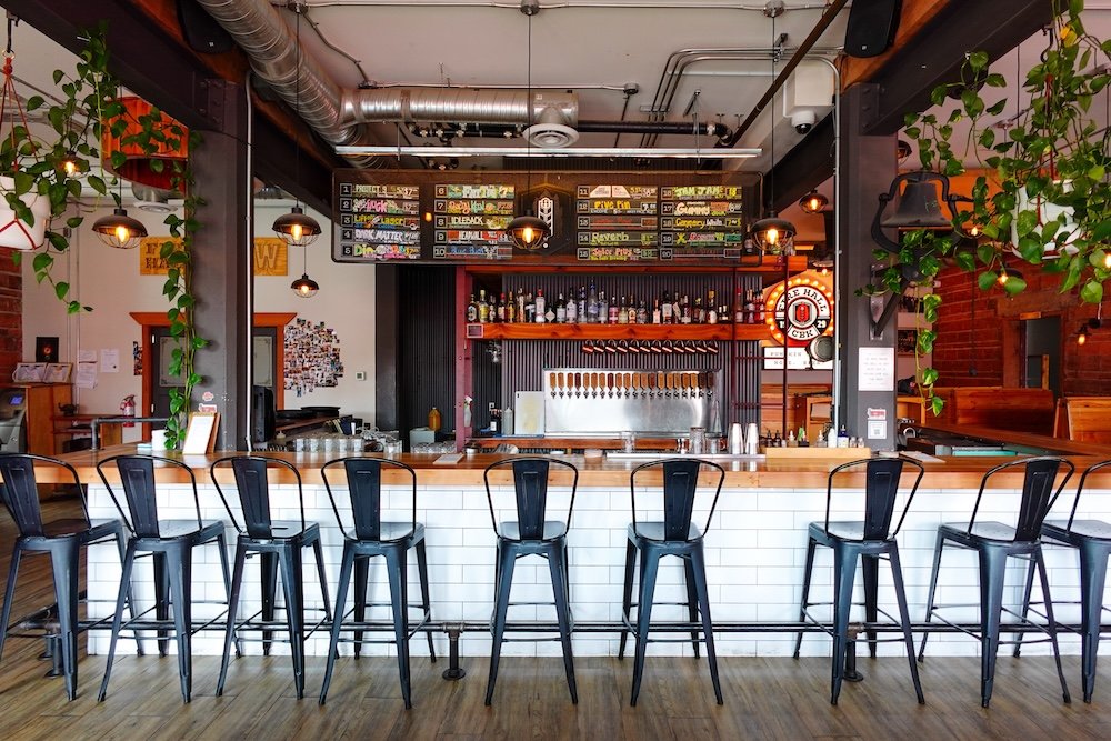 Cranbrook, BC at The Fire Hall Kitchen & Tap shows the stylish bar interior with high stools, exposed brick, hanging plants, and a large chalkboard beer menu highlighting local craft brews in a converted historic fire hall setting.
