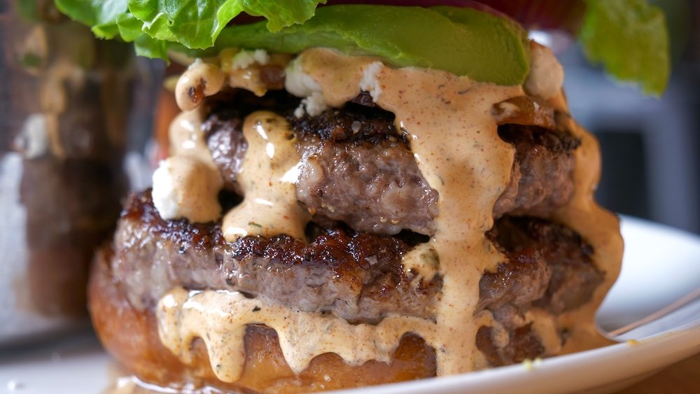 Cranbrook, British Columbia close-up of a double-patty gourmet burger at Fire Hall Kitchen & Tap, stacked with goat cheese, avocado, crispy bacon, and house sauce, showcasing the indulgent, over-the-top burger style the restaurant is known for.
