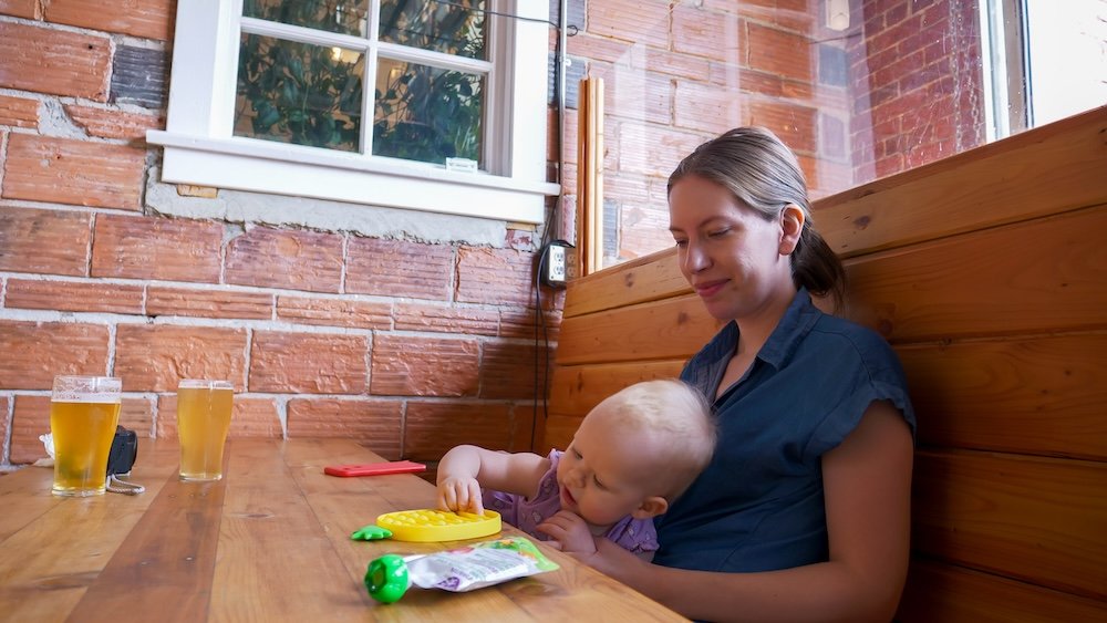 Cranbrook, British Columbia family-friendly dining at Fire Hall Kitchen and Tap, showing baby Aurelia happily playing with toys beside Audrey Bergner at a wooden booth, highlighting the relaxed, welcoming atmosphere for parents traveling with young children.
