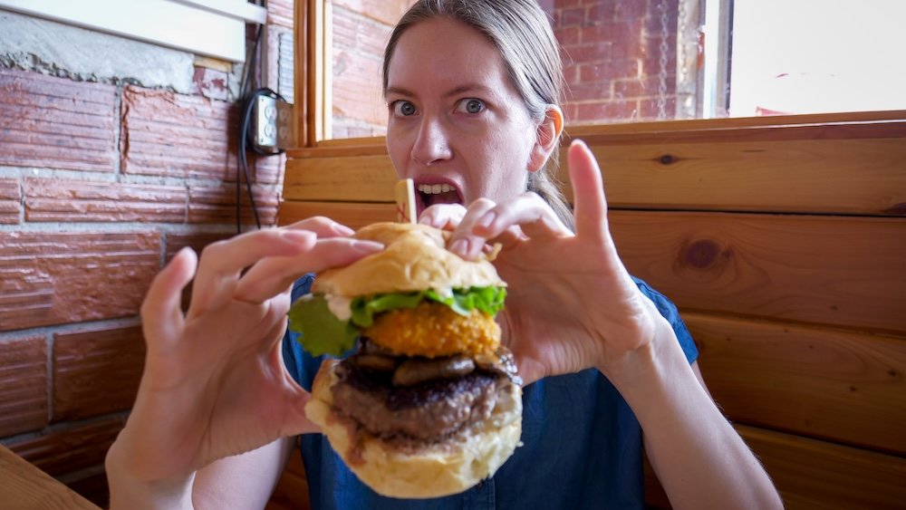 Cranbrook, British Columbia gourmet burger dining at Fire Hall Kitchen & Tap, where Audrey Bergner reacts mid-bite to an over-the-top stacked burger, capturing the playful, indulgent experience of eating one of the restaurant’s signature burgers inside the historic fire hall.
