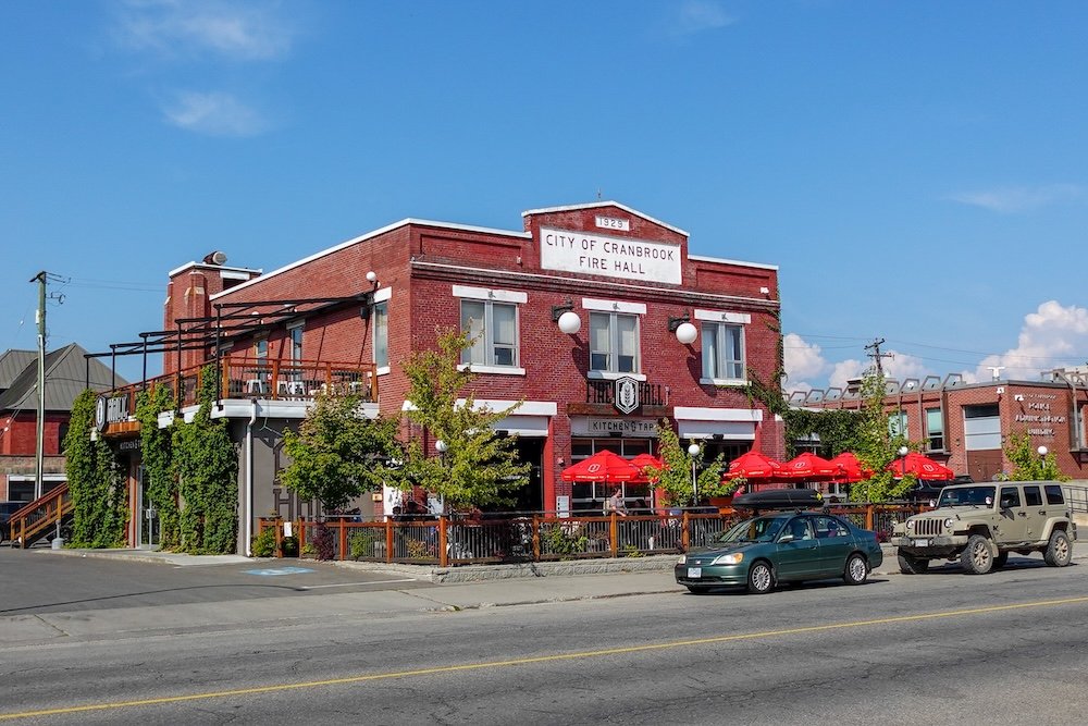 Cranbrook, British Columbia Fire Hall building in summer, now home to Fire Hall Kitchen & Tap, a restored red-brick historic fire station with patio umbrellas and greenery, showing how the city blends heritage architecture with local food and drink culture.