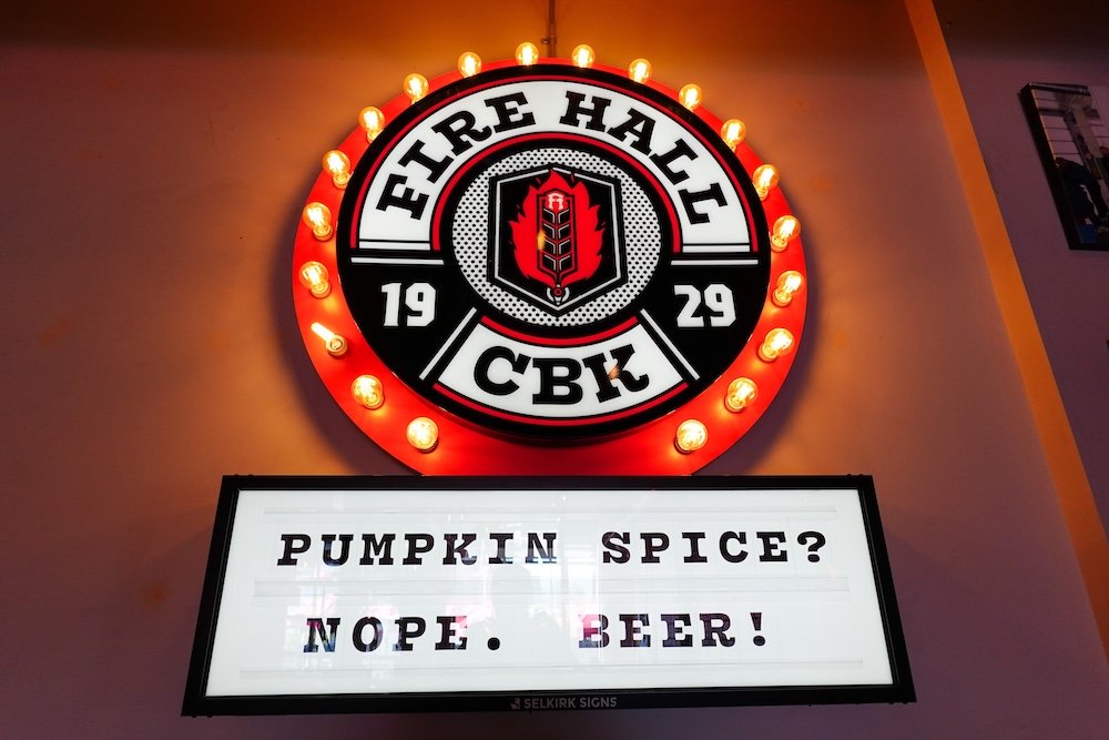 Cranbrook, British Columbia interior signage at Fire Hall Kitchen and Tap featuring the iconic illuminated Fire Hall 1929 logo above a playful “Pumpkin Spice? Nope. Beer!” sign, capturing the restaurant’s humor, craft beer focus, and historic fire hall atmosphere.
