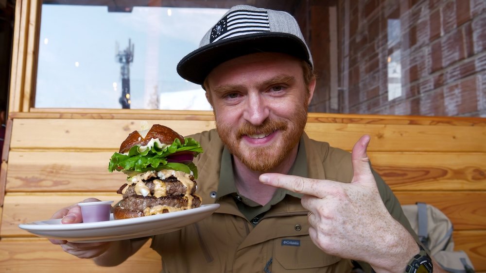 Cranbrook, British Columbia gourmet burger at Fire Hall Kitchen & Tap, with Nomadic Samuel smiling and pointing at a towering double-patty burger inside the historic firehall restaurant, highlighting one of the best places to eat in town.