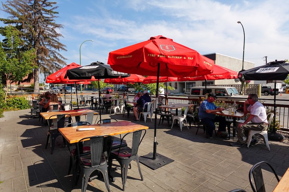 Cranbrook, BC patio dining scene at Fire Hall Kitchen & Tap in early September, showing outdoor tables under red umbrellas during late summer, highlighting why shoulder season is a great time to visit for warm weather without peak crowds.
