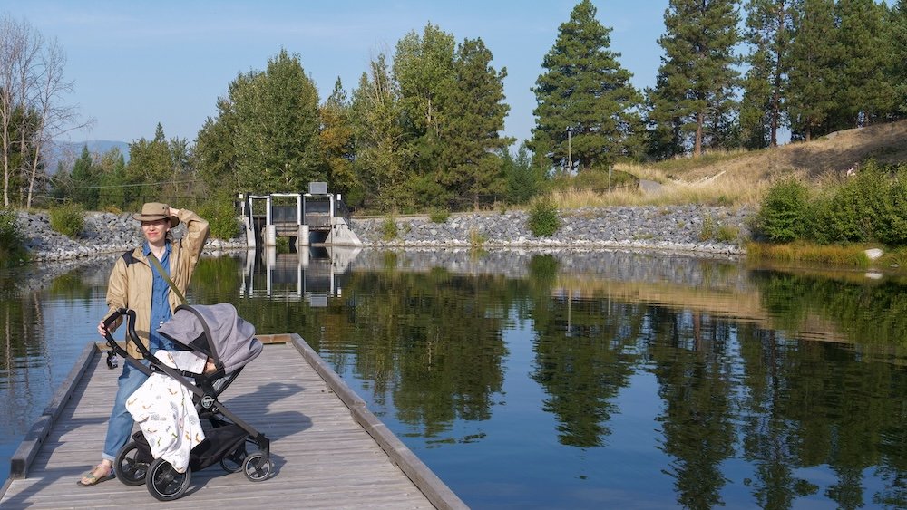 Cranbrook, British Columbia: Idlewild Park lakeside boardwalk with Audrey Bergner strolling with baby Aurelia in a stroller, enjoying calm water reflections, trees, and an easy family-friendly walk in one of Cranbrook’s most relaxing local parks.