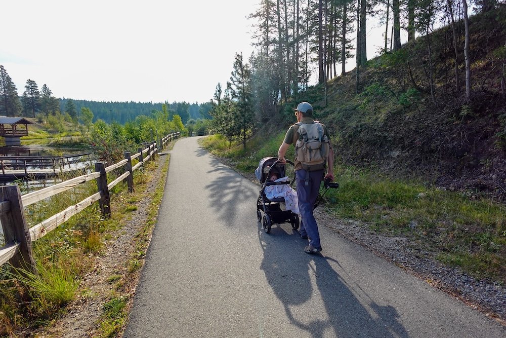 Cranbrook, British Columbia: Nomadic Samuel pushes baby Aurelia in a stroller along the paved trail at Idlewild Park, enjoying a peaceful family walk surrounded by pine trees, wetlands, and gentle hills on a relaxed East Kootenays weekend.