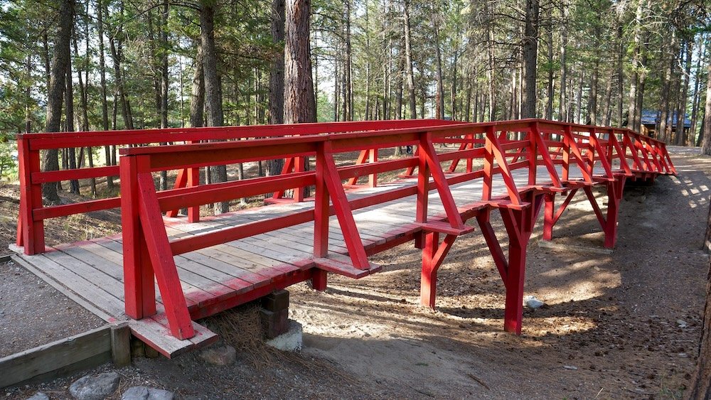 Cranbrook, BC park landmark showing the bright red pedestrian bridge at Idlewild Park, surrounded by tall pine trees and packed dirt trails, highlighting one of the most recognizable features along this peaceful walking loop.
