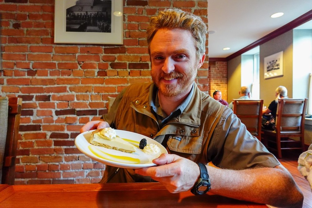 Cranbrook, British Columbia dessert at Numa Lounge & Dining at St. Eugene Resort, with Nomadic Samuel smiling and holding a second dessert plate inside the historic brick-walled dining room during a relaxed and indulgent evening meal.