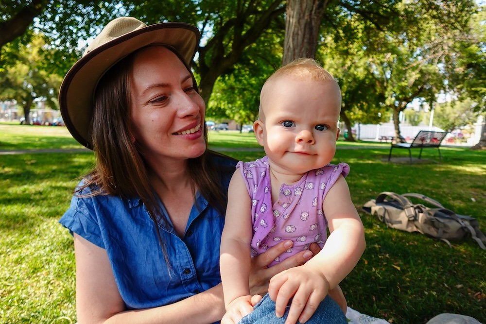 Elizabeth Lake? No → Rotary Park, Cranbrook, BC, with Audrey Bergner holding baby Aurelia while relaxing in the shade of leafy trees, capturing a peaceful family travel moment during a sunny day exploring Cranbrook’s green spaces together.
