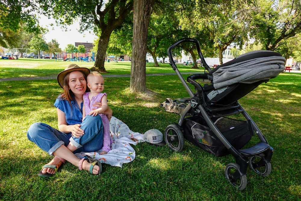 Rotary Park in Cranbrook, British Columbia on a sunny summer day, where green lawns and shady trees frame a relaxed family moment as Audrey Bergner sits on a picnic blanket with baby Aurelia, enjoying an easy, stroller-friendly city park.
