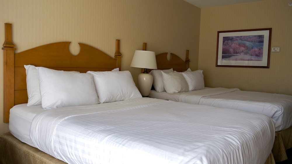 St. Eugene Golf Resort in Cranbrook, BC, showing a comfortable guest room with two neatly made queen beds, warm wooden headboards, soft lighting, and neutral décor that highlights the cozy and restful atmosphere of this historic resort stay.