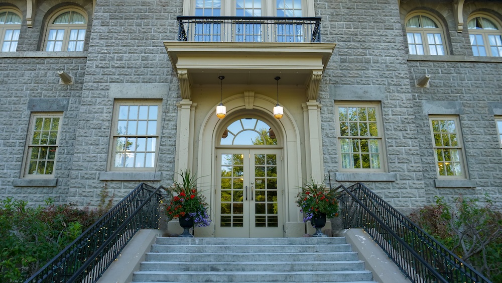 St. Eugene Golf Resort near Cranbrook, BC, showing the historic stone mission building entrance with arched doorway, hanging lantern lights, balcony above, and flower planters framing the staircase, highlighting the preserved heritage architecture of this resort stay.