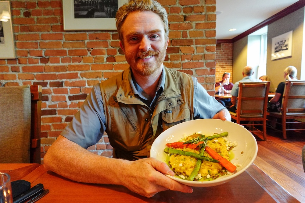 St. Eugene Golf Resort in Cranbrook, BC, with Nomadic Samuel enjoying a plated restaurant meal featuring fresh vegetables and risotto inside the historic mission dining room, showcasing the resort’s cozy atmosphere and on-site dining experience.