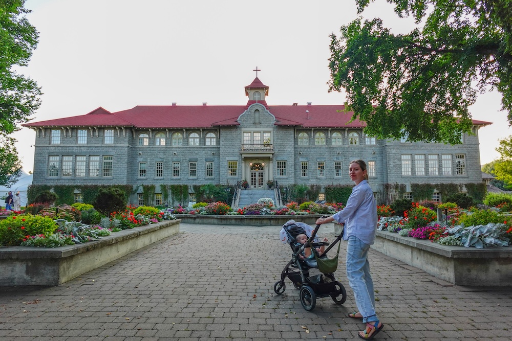 St. Eugene Golf Resort in Cranbrook, BC, with Audrey Bergner pushing a stroller carrying baby Aurelia along the flower-lined pathway toward the historic mission building at sunset, capturing a peaceful family moment at this scenic resort stay.