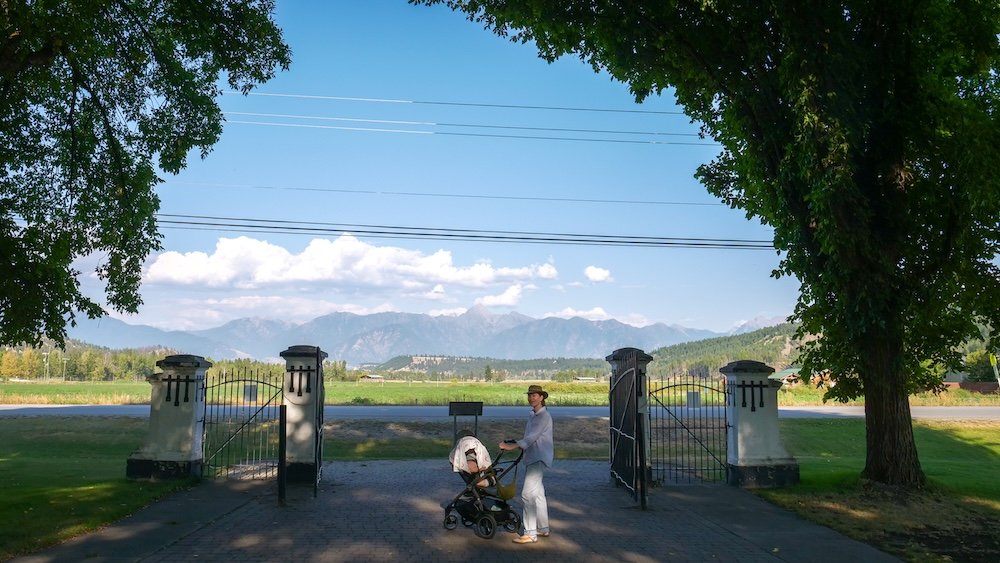 St. Eugene Mission near Cranbrook, BC shows Audrey Bergner strolling with baby Aurelia in a stroller beneath the historic stone gates, with mountain views beyond, capturing a peaceful family visit to this culturally significant landmark in the East Kootenays.
