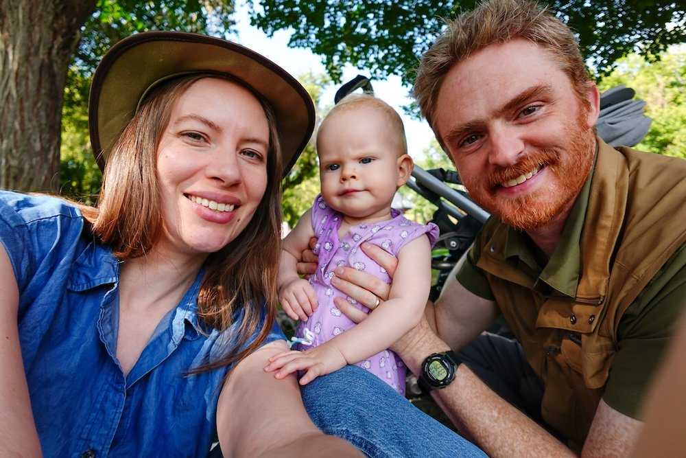 Family park moment in Cranbrook BC A sunny afternoon in Cranbrook, British Columbia, with Audrey Bergner of That Backpacker, Nomadic Samuel Jeffery, and baby Aurelia sitting together in a leafy park, smiling at the camera as Aurelia balances between them near their stroller.