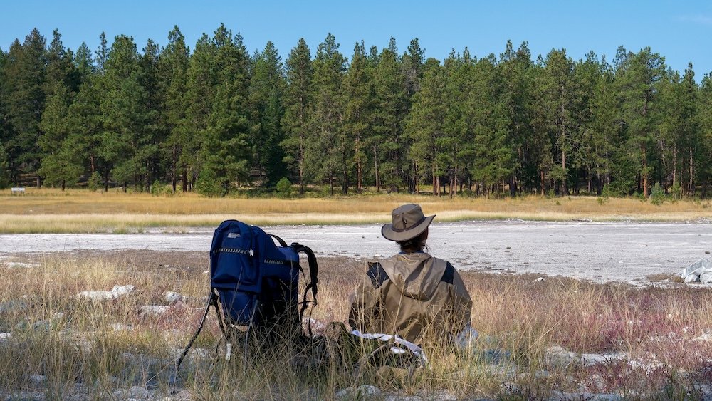 Cranbrook Community Forest scenic viewpoint with Audrey Bergner resting beside a baby carrier, taking in wide-open meadows, dry lakebed terrain, and pine forest backdrops, showing how relaxed breaks and practical gear make family hikes here calm, flexible, and enjoyable near Cranbrook, British Columbia.