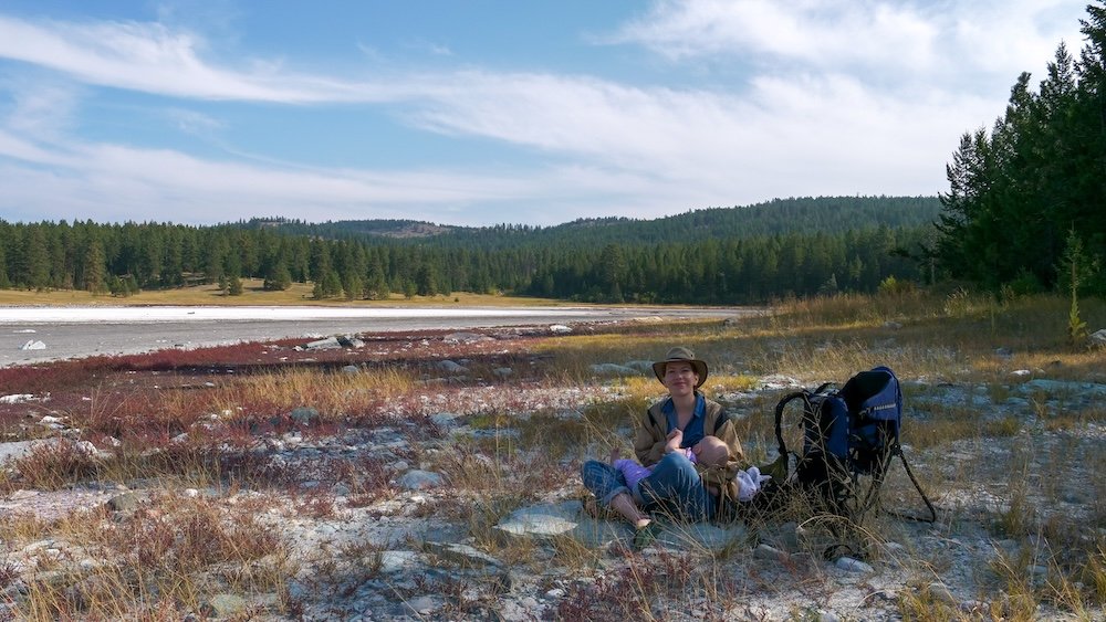 Cranbrook Community Forest Sylvan Lake rest stop with Audrey Bergner sitting on rocky ground while holding baby Aurelia, capturing a peaceful family hiking break surrounded by dry lakebed textures, open meadows, pine forests, and the quiet nature that defines easy hiking near Cranbrook, British Columbia.