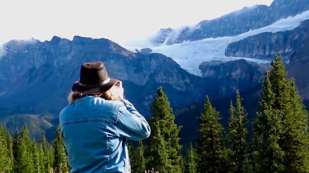 Samuel Jeffery en el mirador del glaciar Crowfoot, Icefields Parkway — Parque Nacional Banff, Alberta Samuel Jeffery fotografiando el glaciar Crowfoot desde el mirador al costado de la ruta en la Icefields Parkway bajo un cielo azul despejado, árboles de hoja perenne en primer plano, Parque Nacional Banff, Alberta, Canadá.