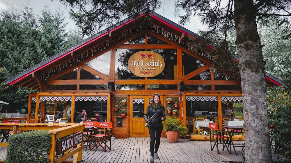 Cucú Schulz eatery, Villa La Angostura, Neuquén Province, Argentine Patagonia Audrey Bergner walks across the wooden deck of Cucú Schulz, an alpine timber eatery in Villa La Angostura, Neuquén Province, Argentine Patagonia, with outdoor seating and pine trees framing the entrance.