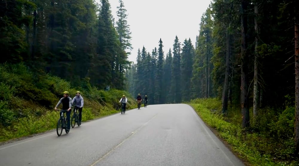 A group of cyclists rides along the paved, forest-lined Moraine Lake Road in Banff National Park, Alberta, Canada, with no personal vehicles in sight.