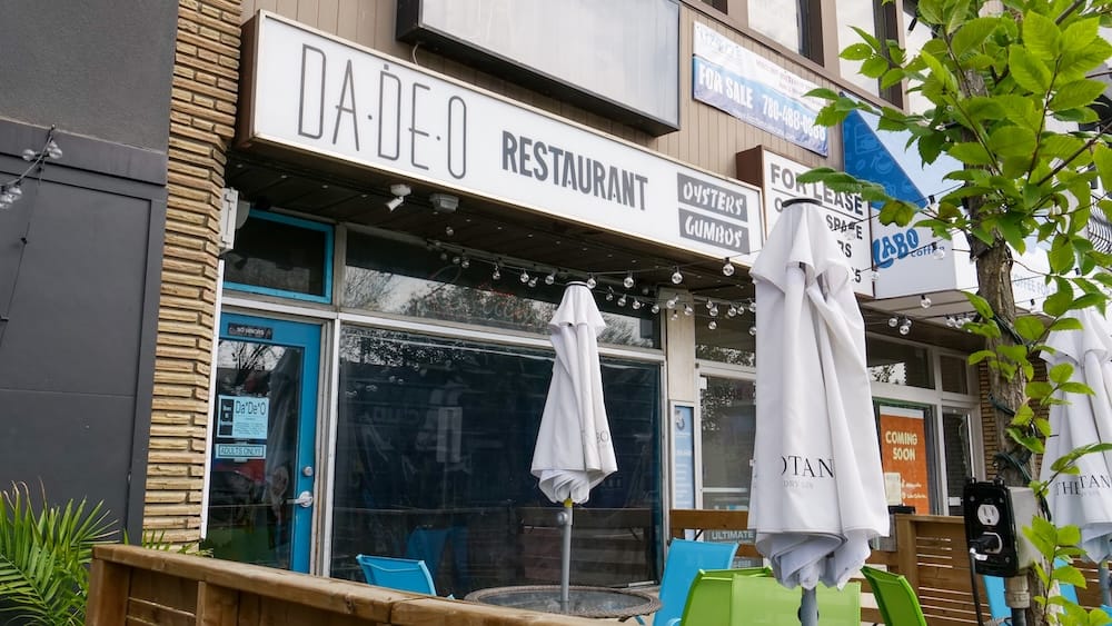 Patio del Restaurante DaDeO, Whyte Avenue, Edmonton, Alberta, Canadá El cartel y la fachada del restaurante DaDeO sobre Whyte Avenue, en Old Strathcona, Edmonton, Alberta, Canadá, con sombrillas plegadas sobre las mesas del patio y luces colgantes arriba de la entrada.