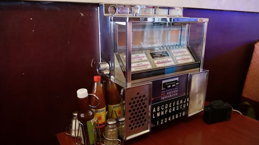Primer Plano de la Rocola de Mesa en DaDeO, Whyte Avenue, Old Strathcona, Edmonton, Alberta, Canadá Primer plano de una rocola de mesa vintage Seeburg Consolette en el restaurante DaDeO en Whyte Avenue en Edmonton, Alberta, Canadá, con tarjetas de selección de canciones que funcionan con monedas de 25 centavos, junto a botellas de salsa picante de Luisiana y condimentos de mesa.