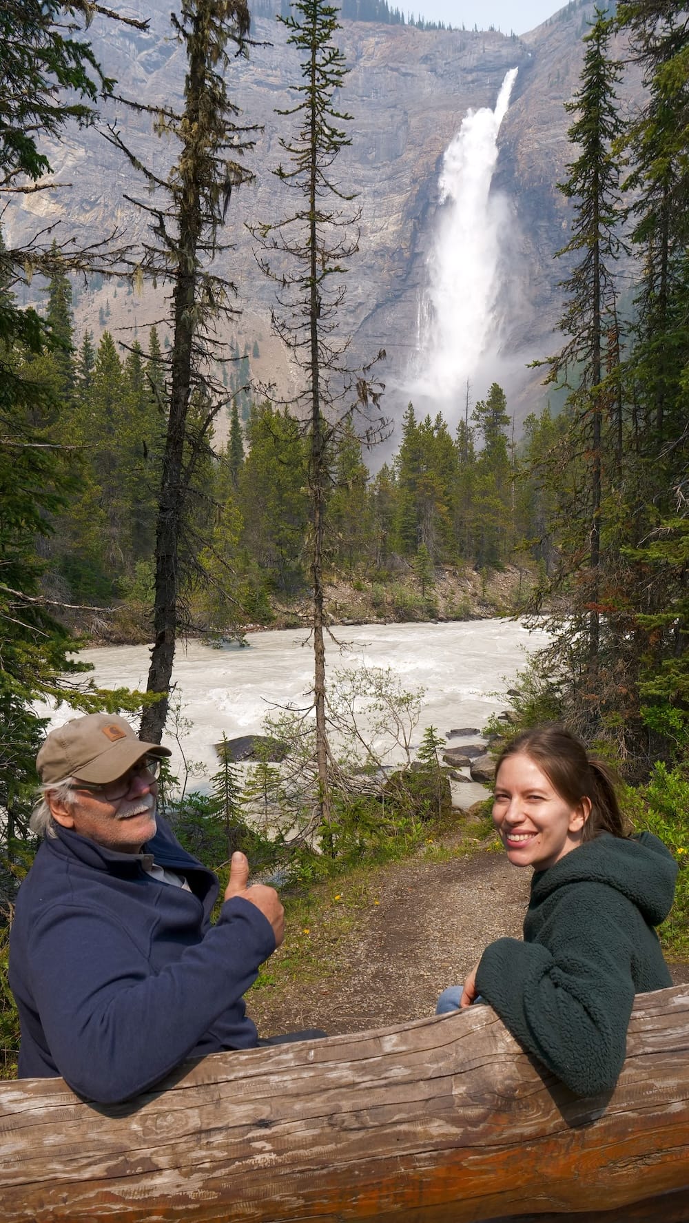 Daniel Bergner gives a thumbs-up while sitting with Audrey Bergner on a wooden railing overlooking the Kicking Horse River and Takakkaw Falls in Yoho National Park, British Columbia, with the waterfall visible through the trees behind them.
