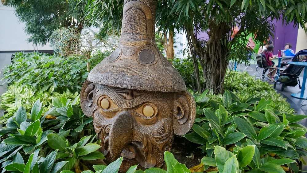 Escultura tallada entre vegetación tropical dentro de Devonian Gardens, Calgary, Alberta, Canadá Una escultura tallada en madera sobresale entre densas plantas tropicales dentro de Devonian Gardens, en Calgary, Alberta, Canadá, rodeada de follaje, árboles y áreas para sentarse en el jardín interior.