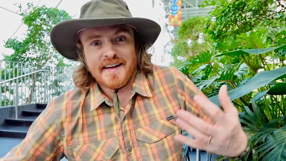 Samuel Jeffery gesticula animadamente junto a vegetación tropical densa dentro de Devonian Gardens en Calgary, Alberta, Canadá, con sombrero de ala ancha y camisa a cuadros.