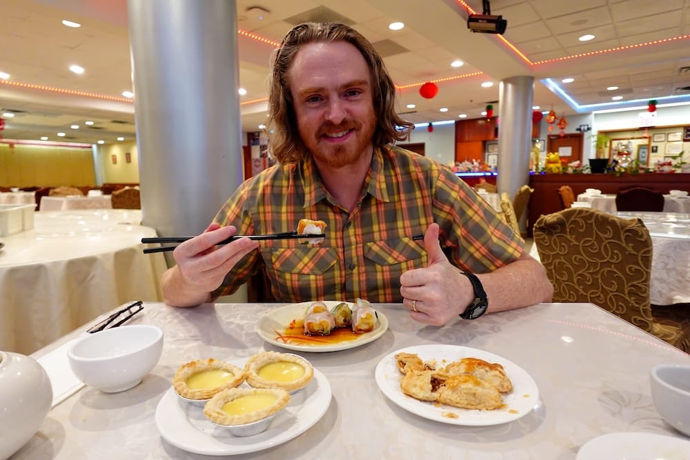 Samuel Jeffery holds chopsticks over a spring roll at a Chinese restaurant table in Calgary, Alberta, Canada, with egg tarts and a baked pastry also plated in front of him.