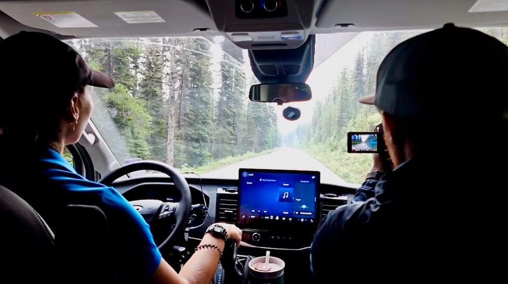 A Discover Banff Tours guide drives a van along Moraine Lake Road in Banff National Park, Alberta, Canada, with Samuel Jeffery filming from the back seat.