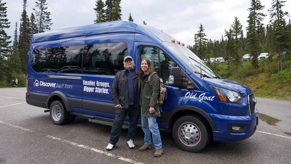 Audrey Bergner and Daniel Bergner standing beside a blue Discover Banff Tours minibus during a Lake Louise outing in Banff National Park, Alberta, Canada, with evergreen forest and parked vehicles behind them.