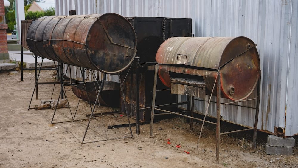 Weathered barbecue drums used for parrilla cooking in Dolavon Patagonia Argentina, sitting beside corrugated metal walls in a quiet rural Welsh-Patagonian village setting.
