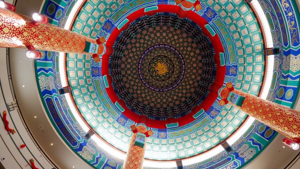 Looking straight up at the ornate domed ceiling of the Chinese Cultural Centre's main hall in Calgary, Alberta, Canada, showing red-and-gold columns and layered blue, green, and gold geometric patterns radiating around the dragon-decorated centre.