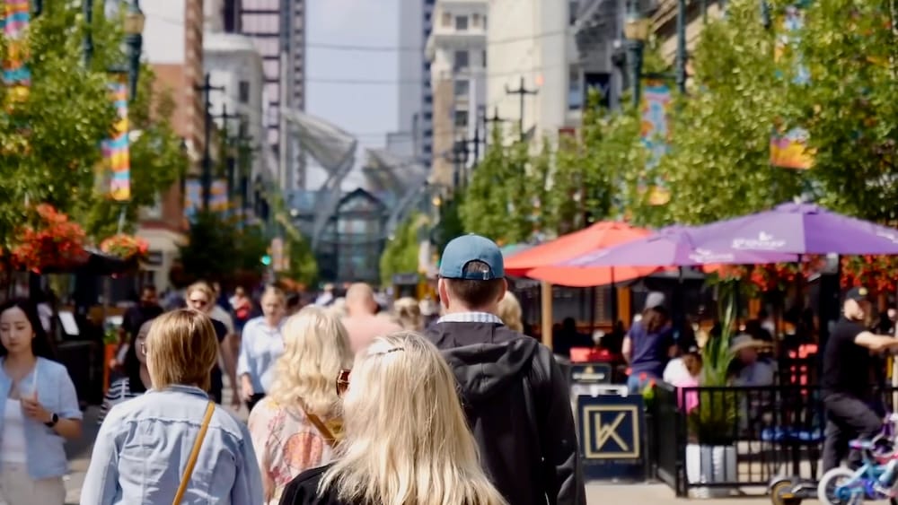 A busy pedestrian street scene in downtown Calgary, Alberta, Canada, shows people walking between leafy trees, outdoor restaurant patios and mid-rise buildings, capturing the city’s lively urban atmosphere.