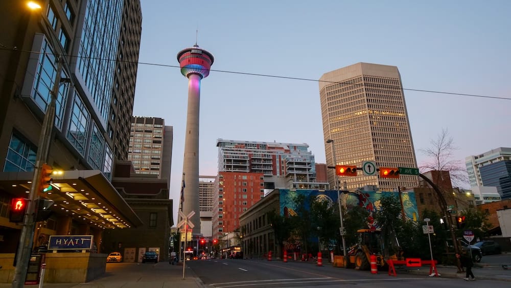 Downtown Calgary, Alberta, Canada at dusk, with the illuminated Calgary Tower rising above office buildings along 7th Avenue South, near a CTrain crossing and a Hyatt hotel entrance.