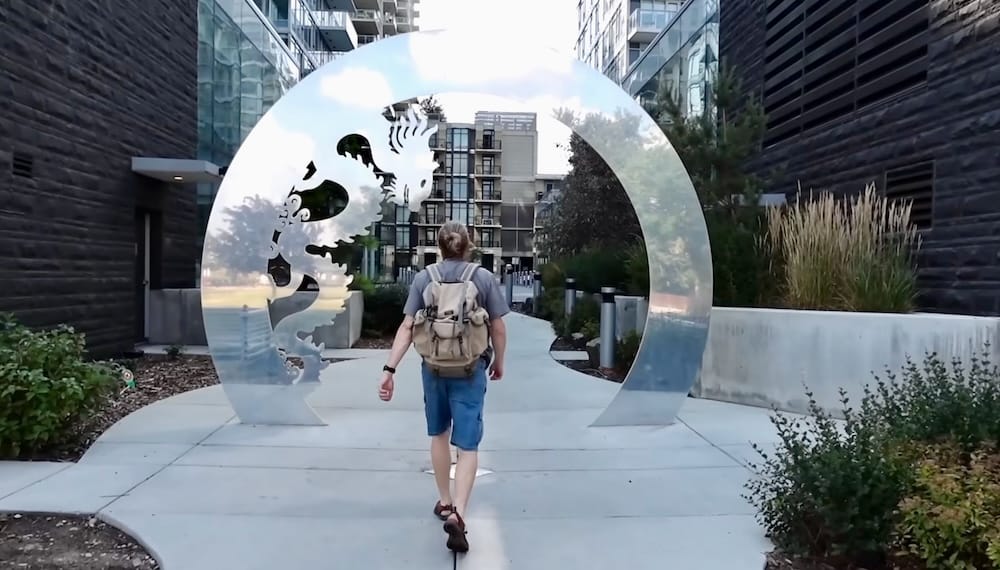 Samuel Jeffery Walking in Downtown Calgary Alberta Canada Beside Public Art on a Car-Free City Route Samuel Jeffery walks through a landscaped pedestrian passage in Downtown Calgary, Alberta, Canada, approaching a large circular metal public-art installation between modern residential buildings.